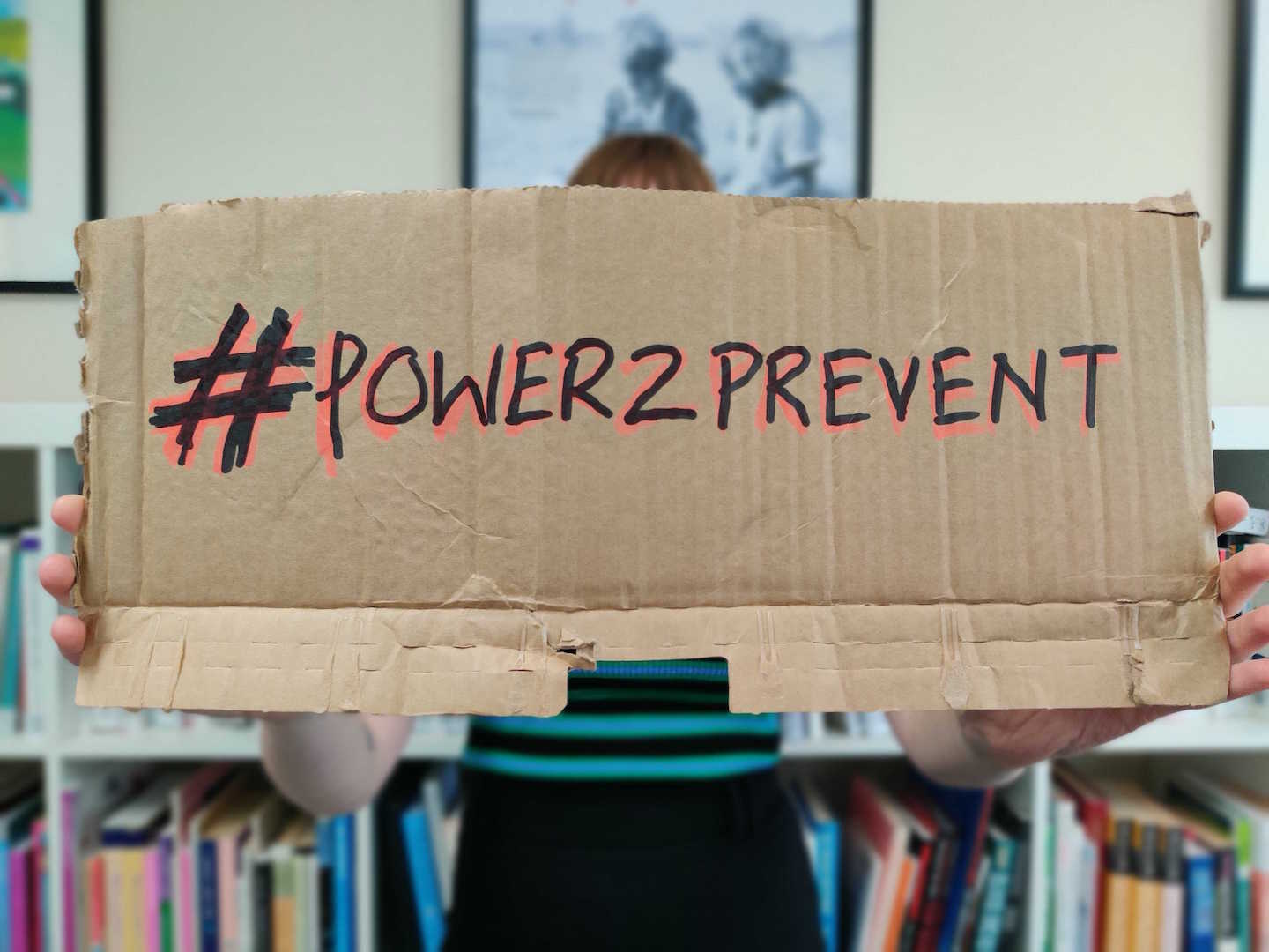 Image: woman holding up cardboard sign with hand written message: #Power2Prevent