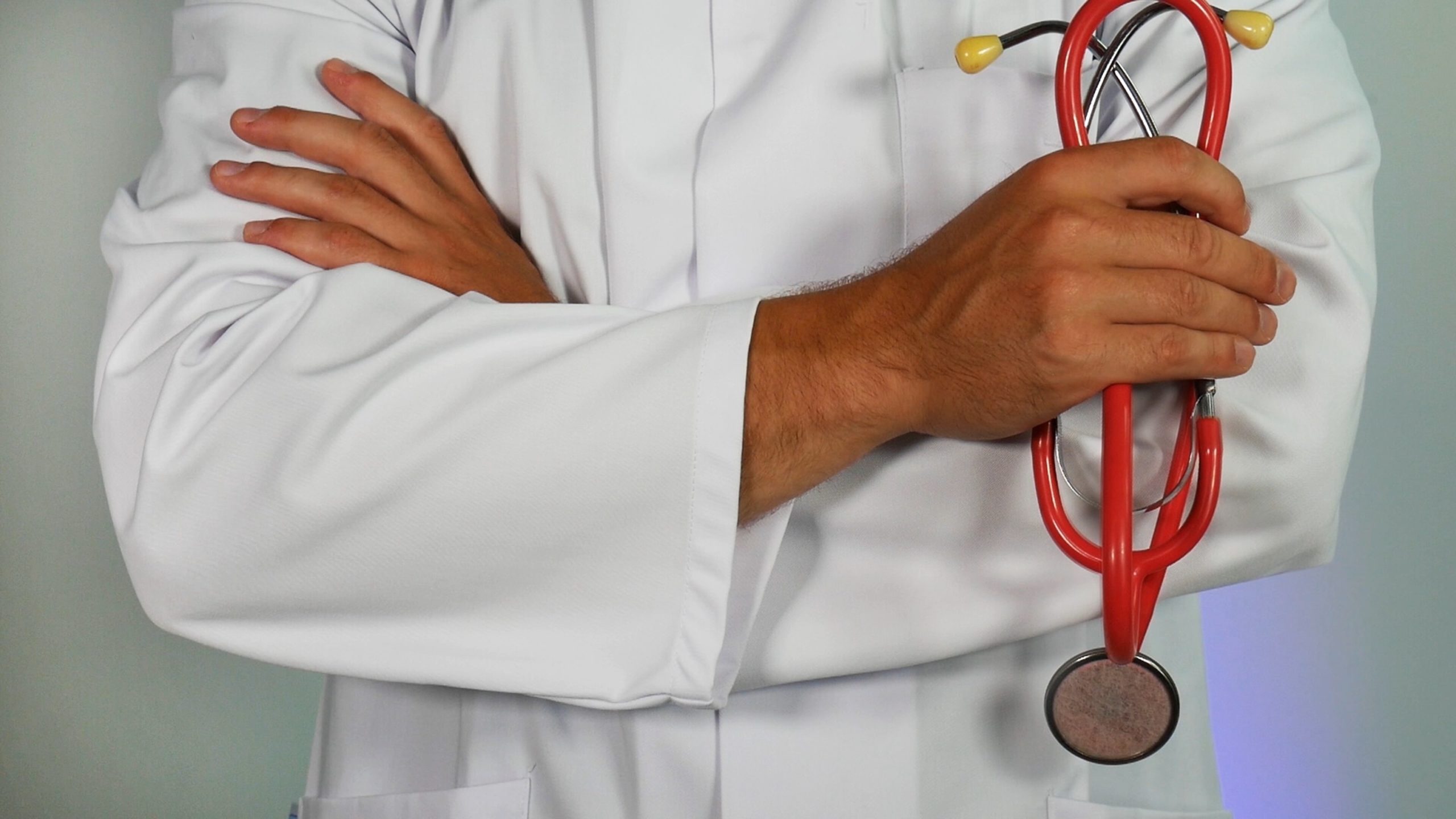 Image: a man standing with his arms cross over his chest. He is wearing a lab coat and holds a stethoscope in his hand.