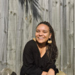 Image: Woman wearing a black jumper and gold earrings, sitting against a garden fence smiling. She has brown skin and long braids.