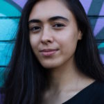 Image: woman with long dark hair and brown skin stands against a bright purple and blue brick wall. 