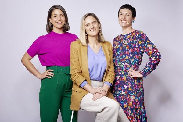 Image: three women are posing closely together. One is blonde, the other brunette. They're all smiling and are wearing bright, happy colours.