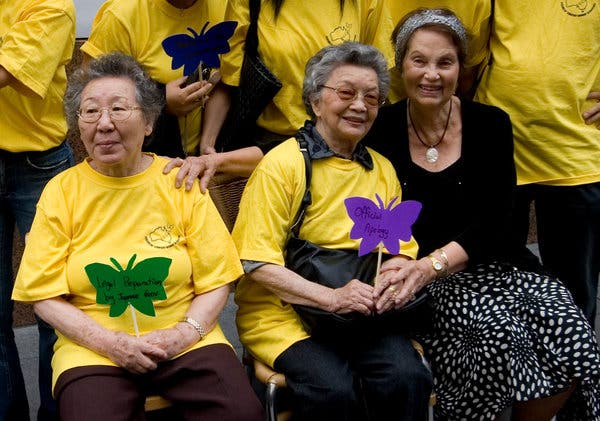 [Image: a group of older women sitting together, holding paper butterflies with the message 'official apology'. Two of the women are Korean, one of the women is white.