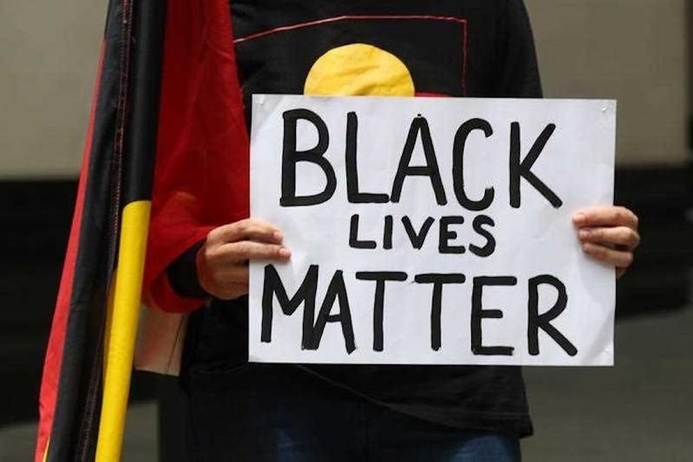 Image: photograph of a person wearing a tshirt with the Aboriginal flag across the front and an Aboriginal flag at their side, holding up a handwritten sign with the following text 'Black Lives Matter'.