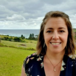 Image description: a woman with short brown hair standing against a background of hills meeting the sea. She's smiling.