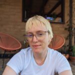 The image shows a woman wearing glasses and smiling. She is sitting near a chair outdoors. Why not on the chair? We'll never know.